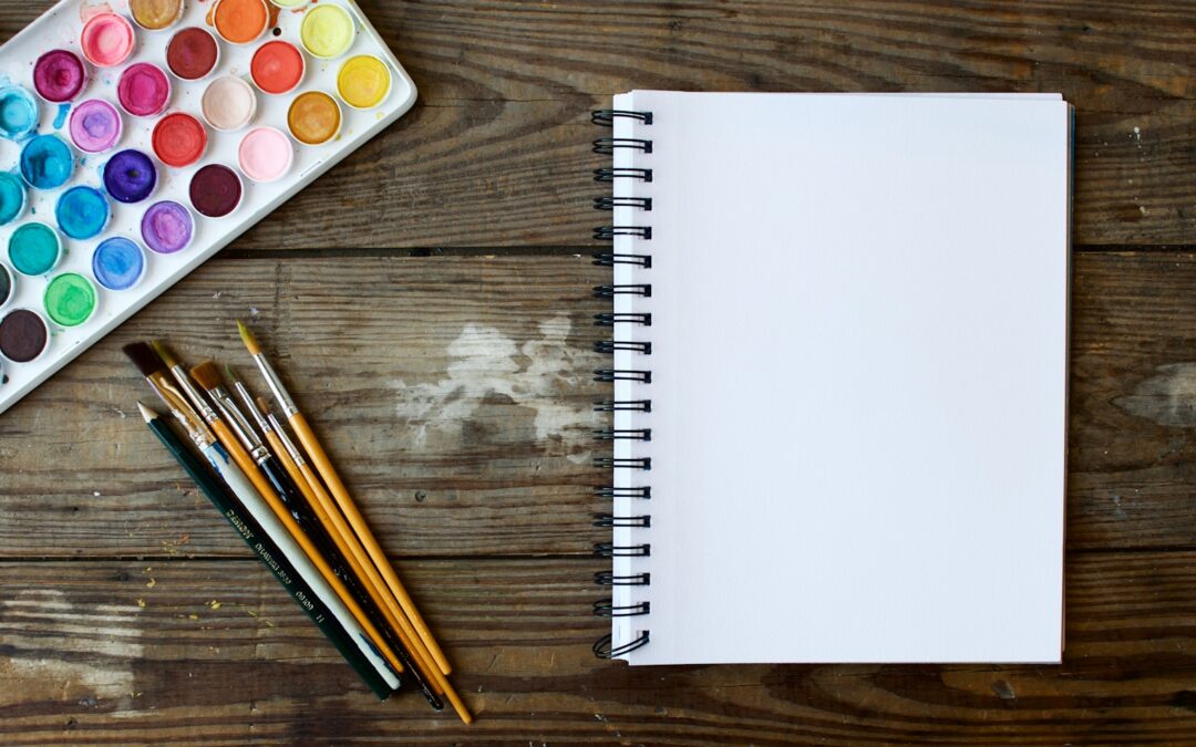 paint brushes next to drawing book and water color palette